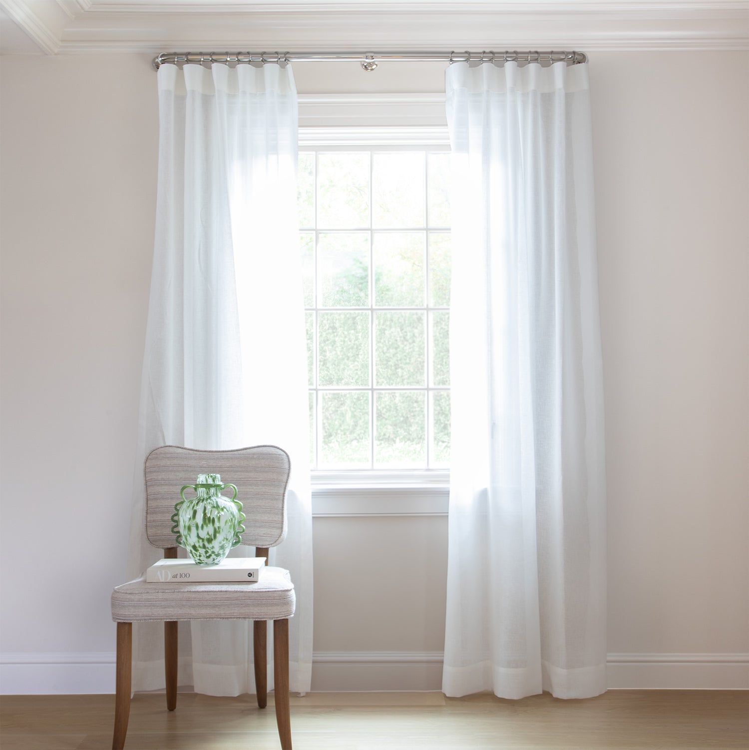 photo of sheer white curtains  hung on a metal rod in front of an illuminated window with a cream chair in front of the window with a cream book and a green and white vase sitting on the chair