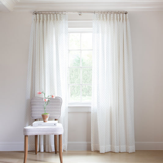 photo of sheer white curtains with embroidered pink polka dots hung on a metal rod in front of an illuminated window with a cream chair in front of the window with a white book and a brown vase with a pink flower sitting on the chair