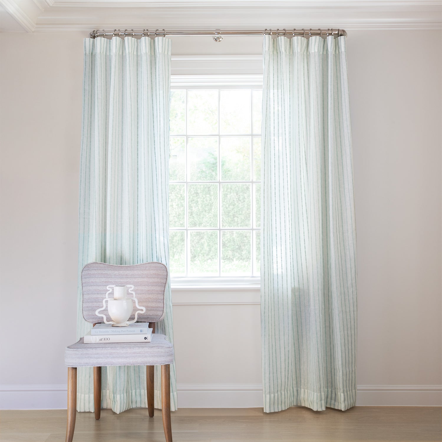 photo of sheer white curtains with embroidered mint green stripe pattern hung on a metal rod in front of an illuminated window with a cream chair in front of the window with two cream books and a cream vase sitting on the chair