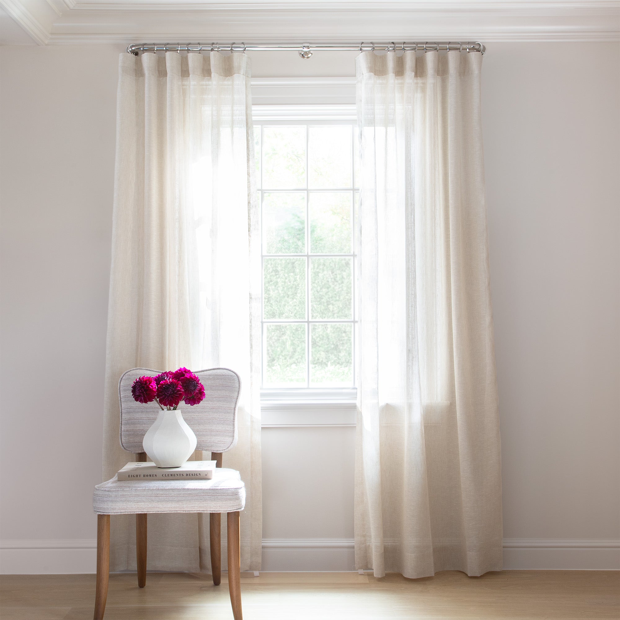 photo of sheer cream curtains  hung on a metal rod in front of an illuminated window with a cream chair in front of the window with a cream book and a white vase with pink flowers in it sitting on the chair