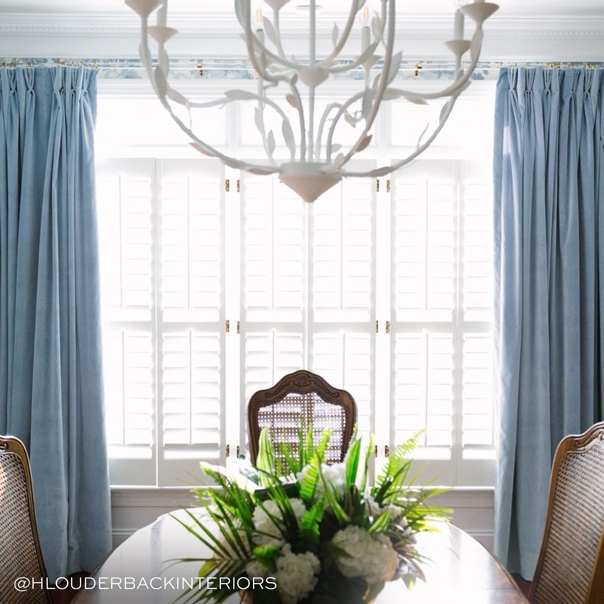 photo of a dining room with a white chandelier hanging, blue velvet curtains hung in front of an illuminated window and a brown wooden table with a floral arrangement on it 