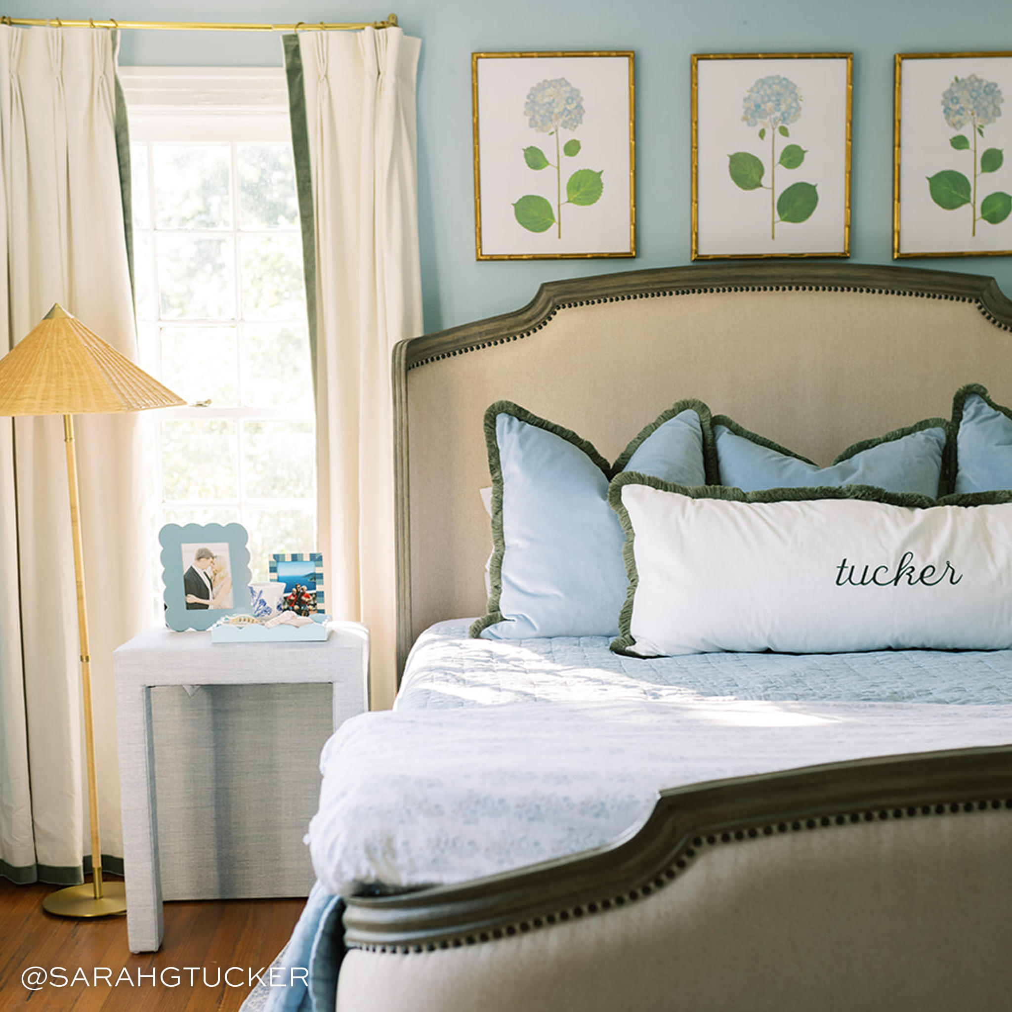 Bedroom with whie curtains with green trim on the window and a cream colored bed in front with three light blue velvet pillows behind a white pillow with embroidery that says "Tucker"