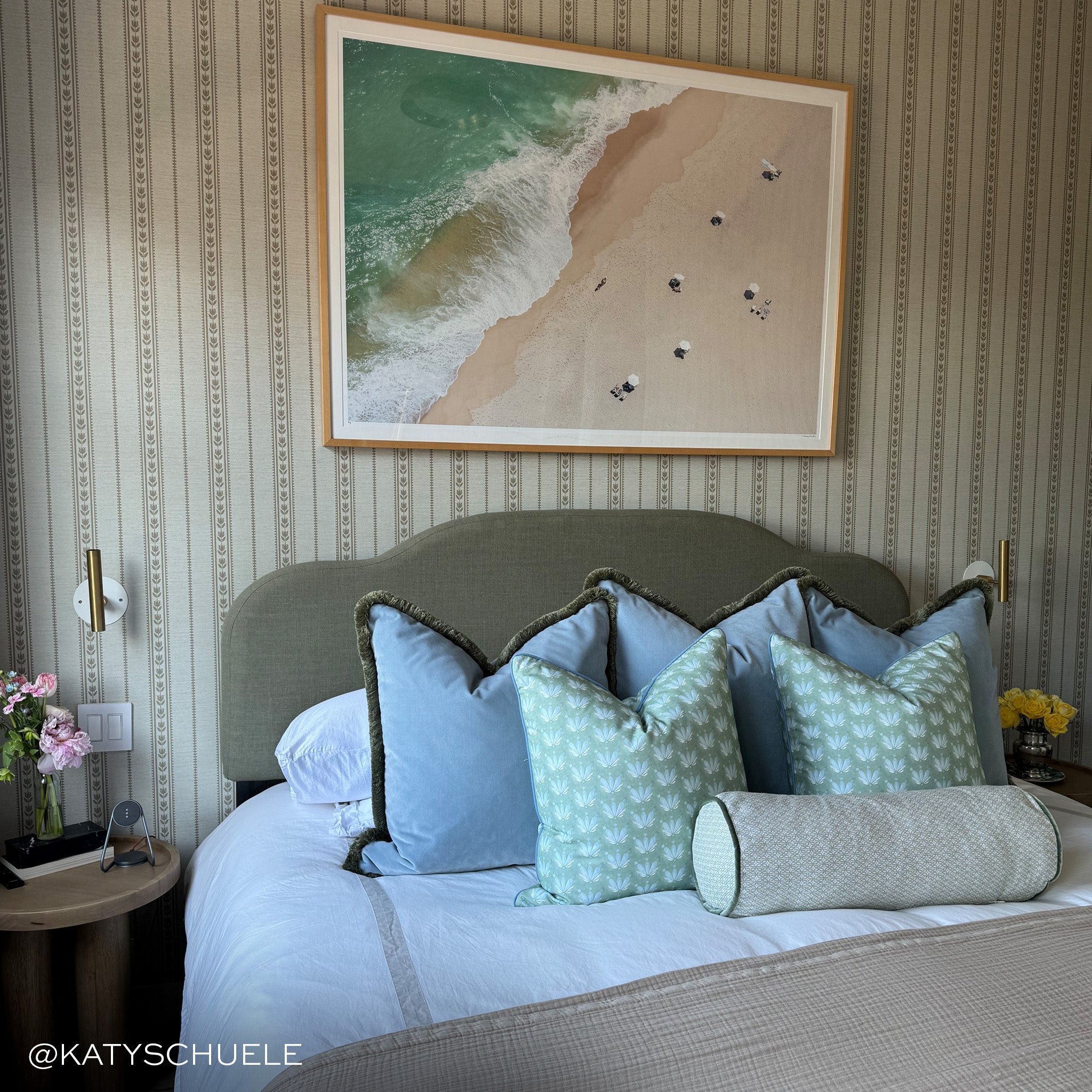 Bedroom with striped wallpaper on the walls and a picture of a beach hung on the wall above the bed with sky blue velvet pillows on the bed