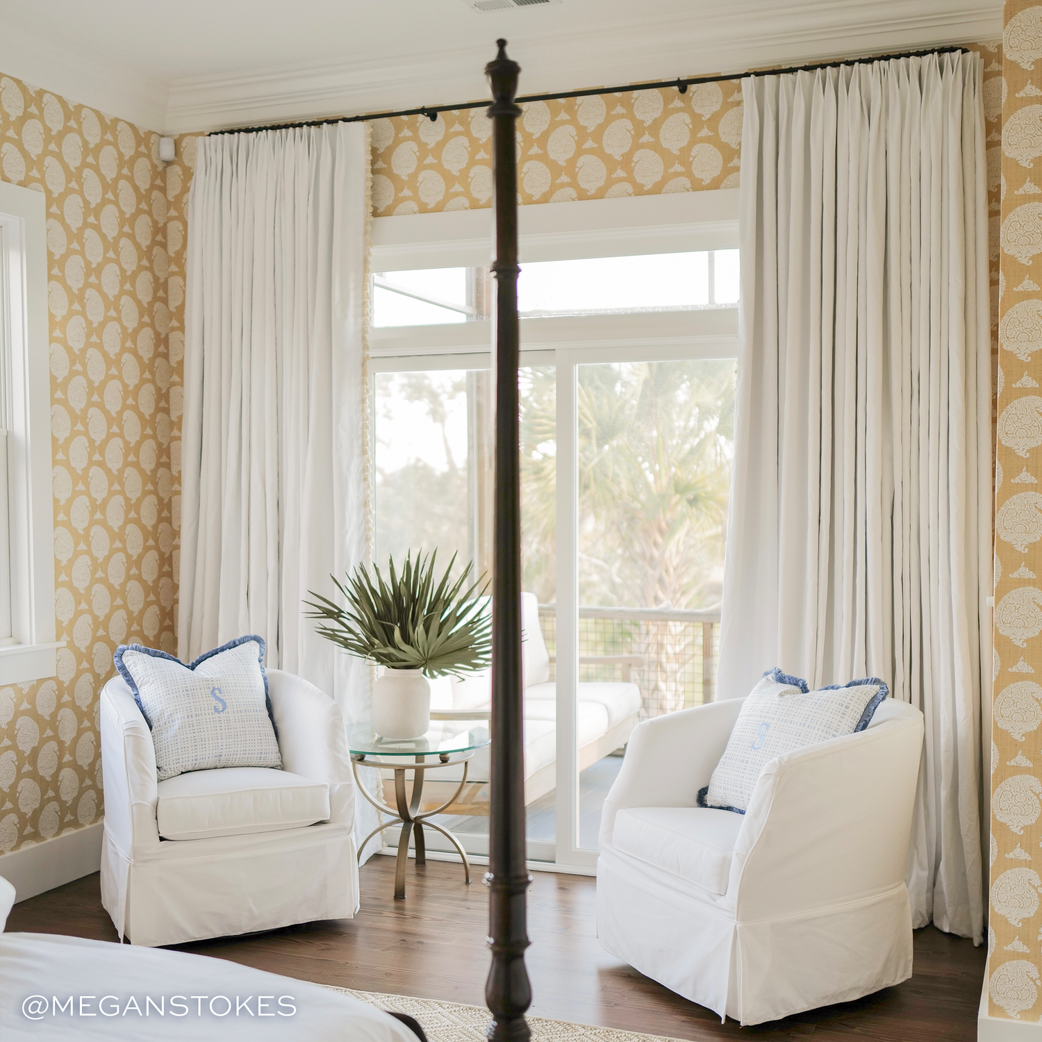a yellow wallpapered room with white cotton curtains hanging from a black rod  behind two white chairs with light blue and white plaid pillows and a green plant between them