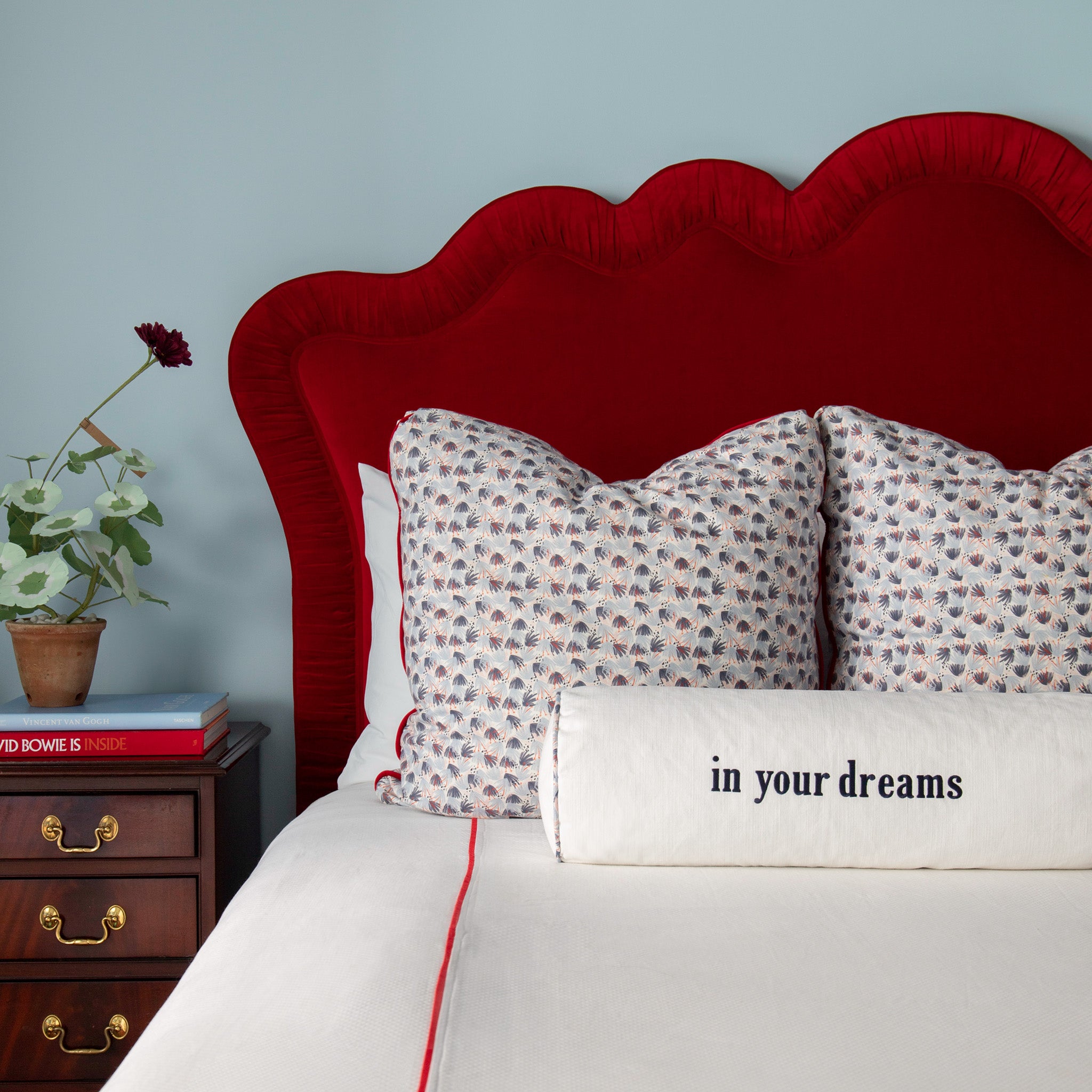 Close-up of bedroom styled with Red and Blue Printed Pillows and white cotton monogrammed bolster on White bed with red headboard next to a wooden nightstand with flowers in vase on top of stacked books
