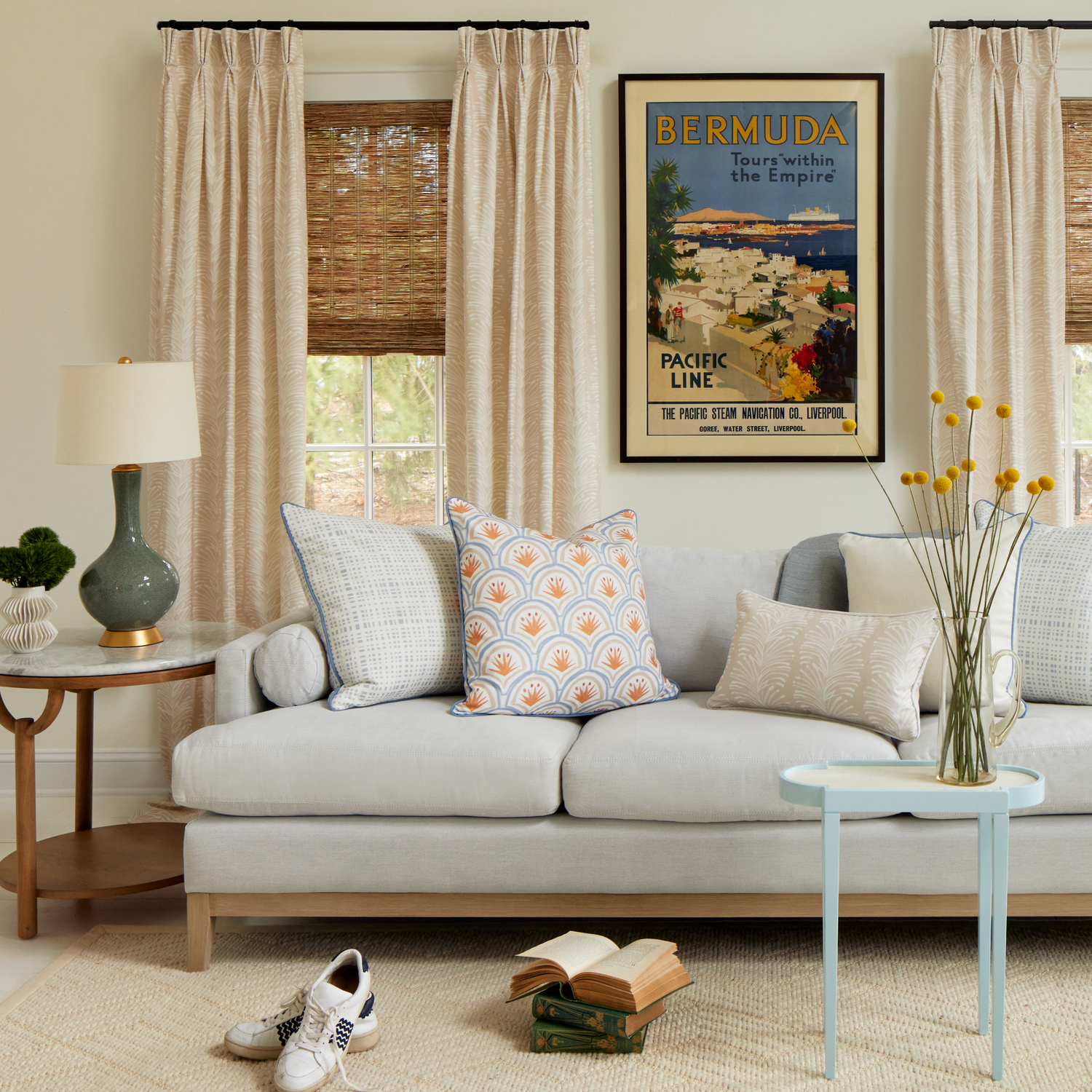 Living room styled with two sky blue gingham printed pillows, Art Deco Palm Pattern Printed Pillow, Beige Botanical Stripe Printed Lumbar, and Light brown Pillow on top of grey couch with Beige Botanical Stripe set of two curtains, books stacked on floor by pair of white tennis shoes and blue and white circular table with yellow flowers in clear vase.