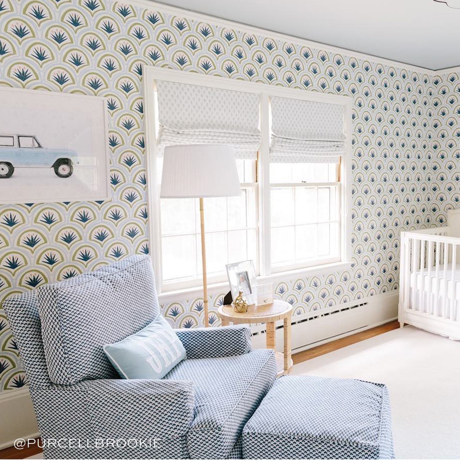 Nursery room styled with Art Deco Palm Pattern Printed Wallpaper by illuminated window next to white sofa chair with monogrammed pillow. Photo taken by Purcell Brookie