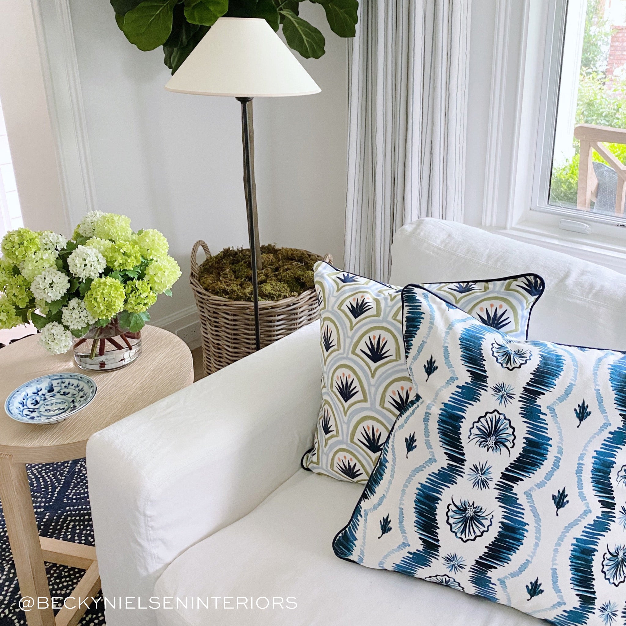 White couch close-up styled with an Art Deco Palm Pattern and a Blue Ikat Stripe Printed pillow next to small circular wooden table with green flowers in clear vase and plate on top. Photo taken by Becky Nielsen Interiors