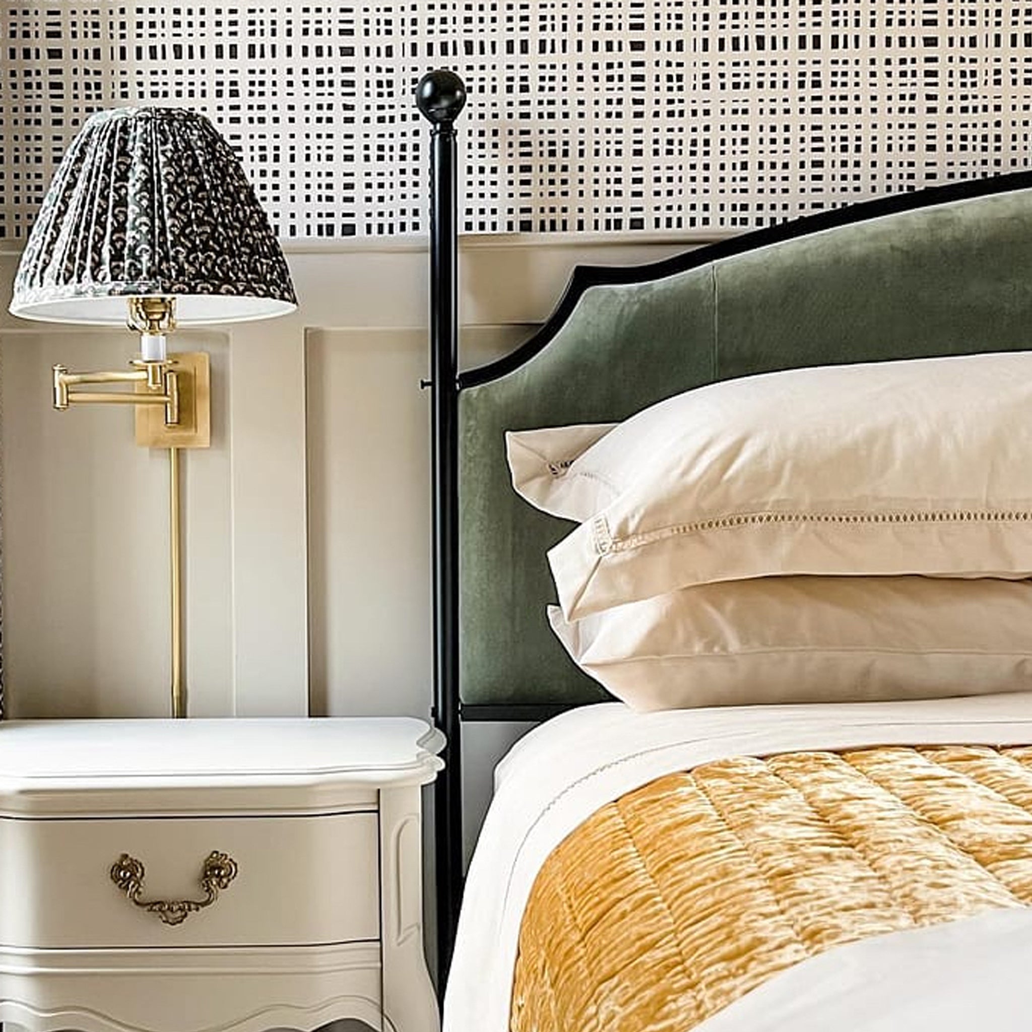 Bedroom corner with upholstered green headboard, beige pillows, quilted mustard blanket, white nightstand, and patterned wall lamp