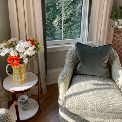 vase of flowers sitting on a white and brown side table with white curtains in front of a window with fern green velvet band and a fern green velvet pillow in a green chair