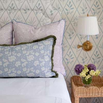 close up of a corner of a bed with white bedding, lavender botanical stripe pillows, and blue floral botanical patterned pillows on the bed with a brown rattan night stand next to the bed with a vase of flowers on it 