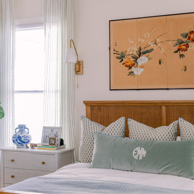 sheer white curtains with embroidered mint green stripes hung in front of an illuminated window with a white side table in front of the window with a blue and white vase on the table and a rattan bed to the right of the table with mint green geometric chenille pillows and a blue green velvet lumbar pillow on the bed