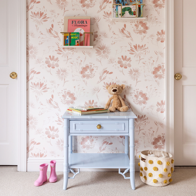 close up of pink floral wallpaper in a bedroom with a light blue table in front of the wall with books and a stuffed animal stacked on top and pink rain boots on the ground next to the table 