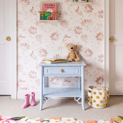 Pink Floral wallpaper in a bedroom with a light blue side table in front of the wall stacked with books and stuffed animals