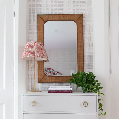 beige plaid print wallpaper on a wall with a mirror hung on the wall and a white dresser in front of it with a green ivy plant in a white pot , lamp with pink lampshade, and books stacked on the dresser