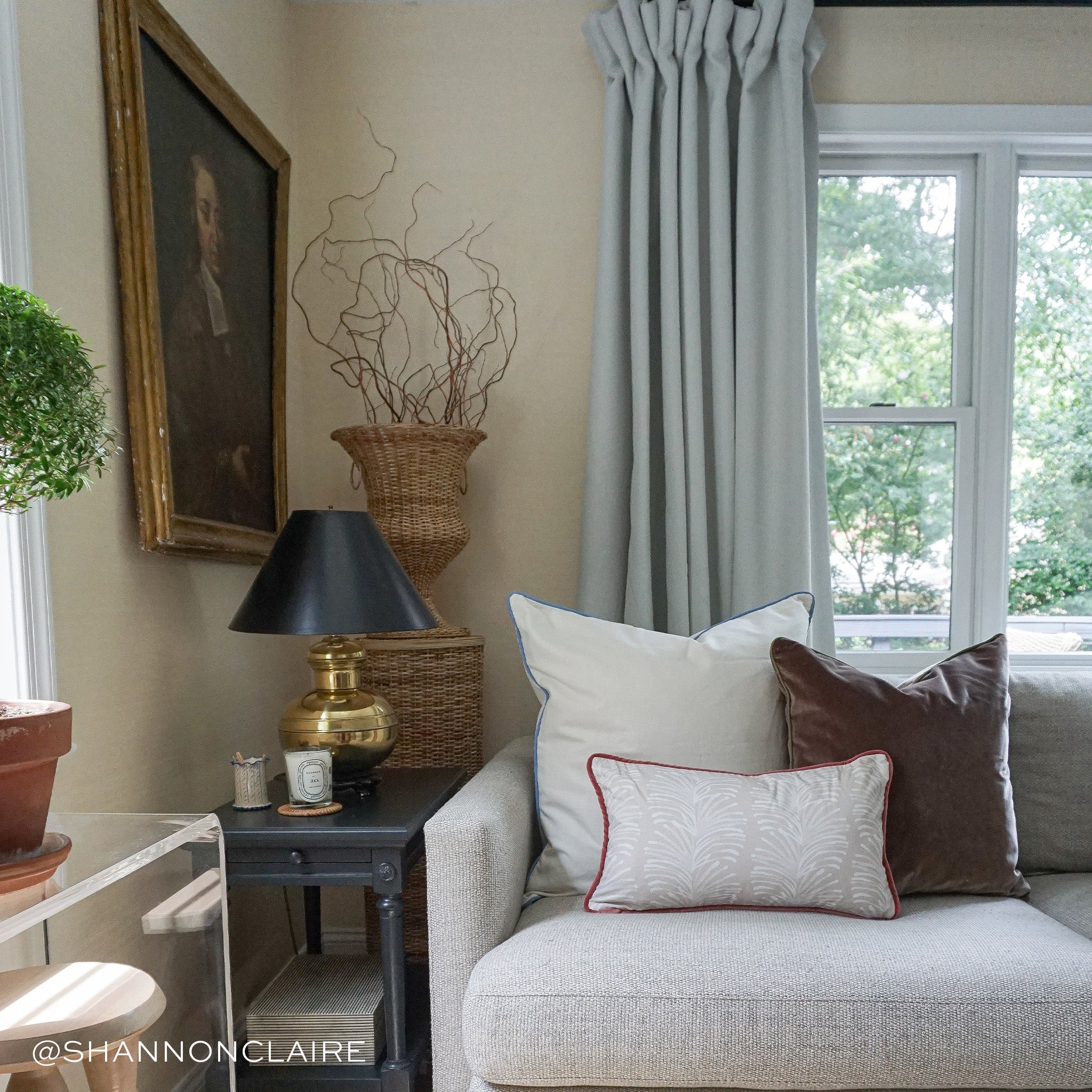 Living room styled with an oat linen pillow, a brown velvet pillow, and a beige palm printed lumbar on grey couch next to black wooden table with black and gold lamp and candle on top next to window with light blue curtains. Photo taken by Shannon Claire