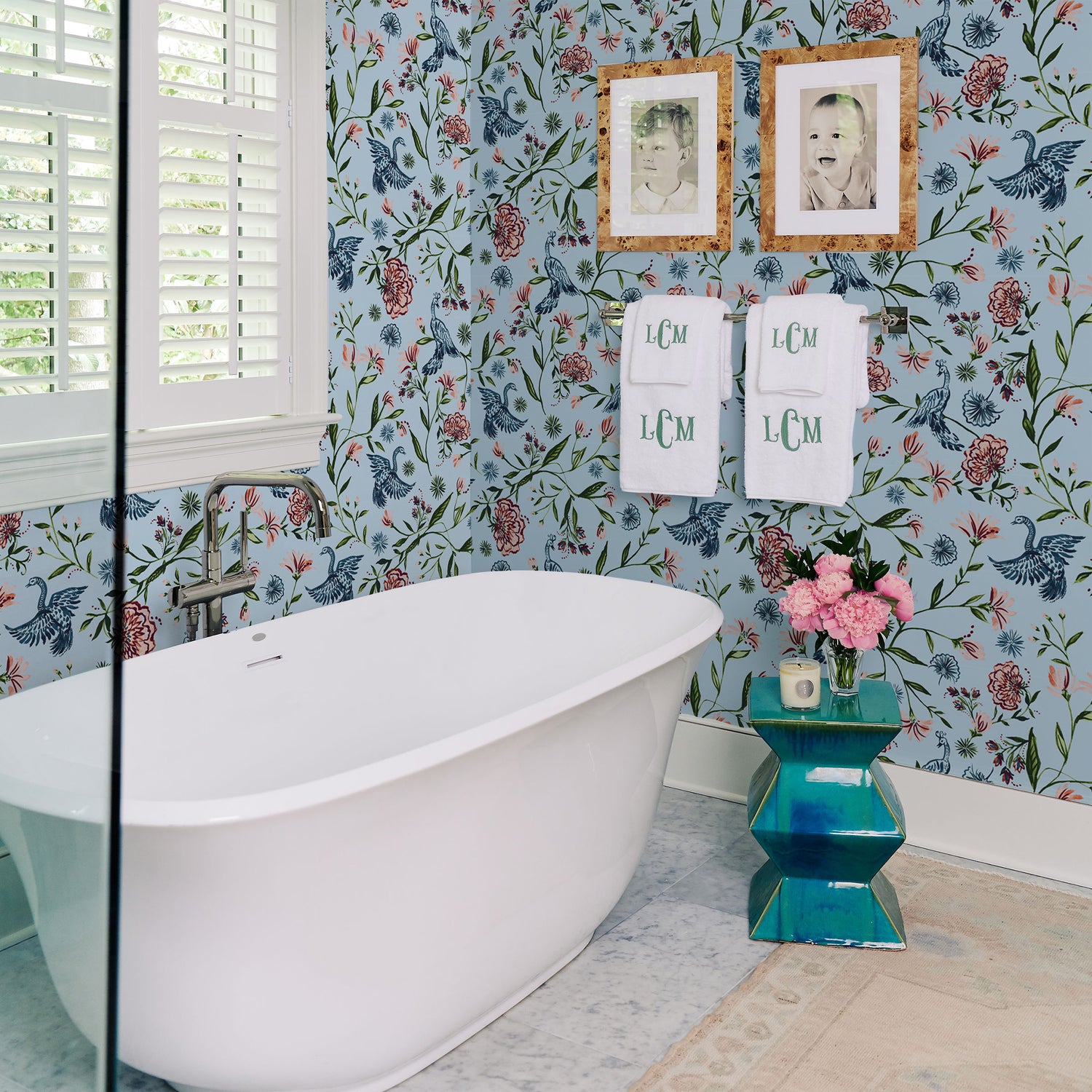 Bathroom styled with Blue Chinoiserie printed wallpaper with white tub in front next to blue metallic small table with pink flowers in vase and candle on top below white monogrammed towels hanging from metal rod and two picture frames on top