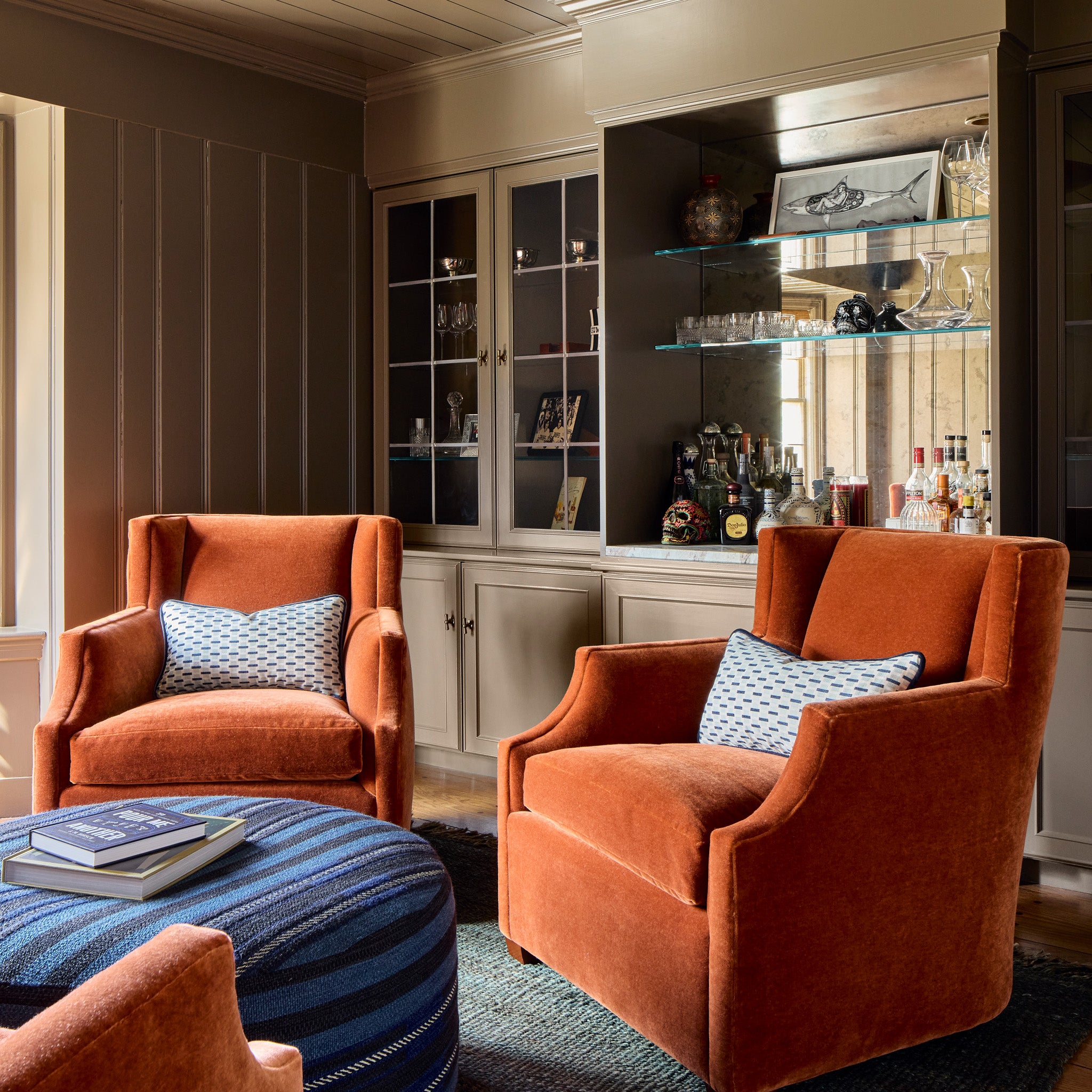 burnt orange chairs with blue geometric pillows on them and a navy blue ottoman stacked with books