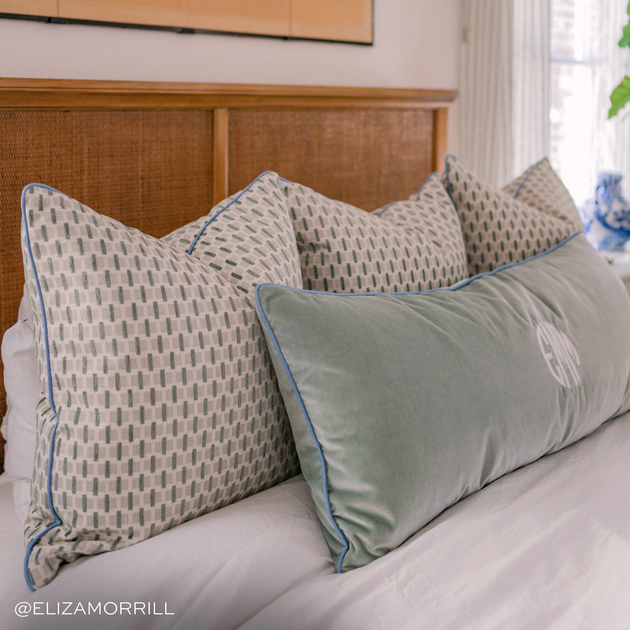 close up photo of a bed with a light brown headboard with mint green chenille jacquard pillows with blue piping trim on the pillows and a sea salt velvet blue green lumbar pillow on the bed with the same blue piping on the pillow and an embroidered monogram on it sitting on the bed with white bedding 