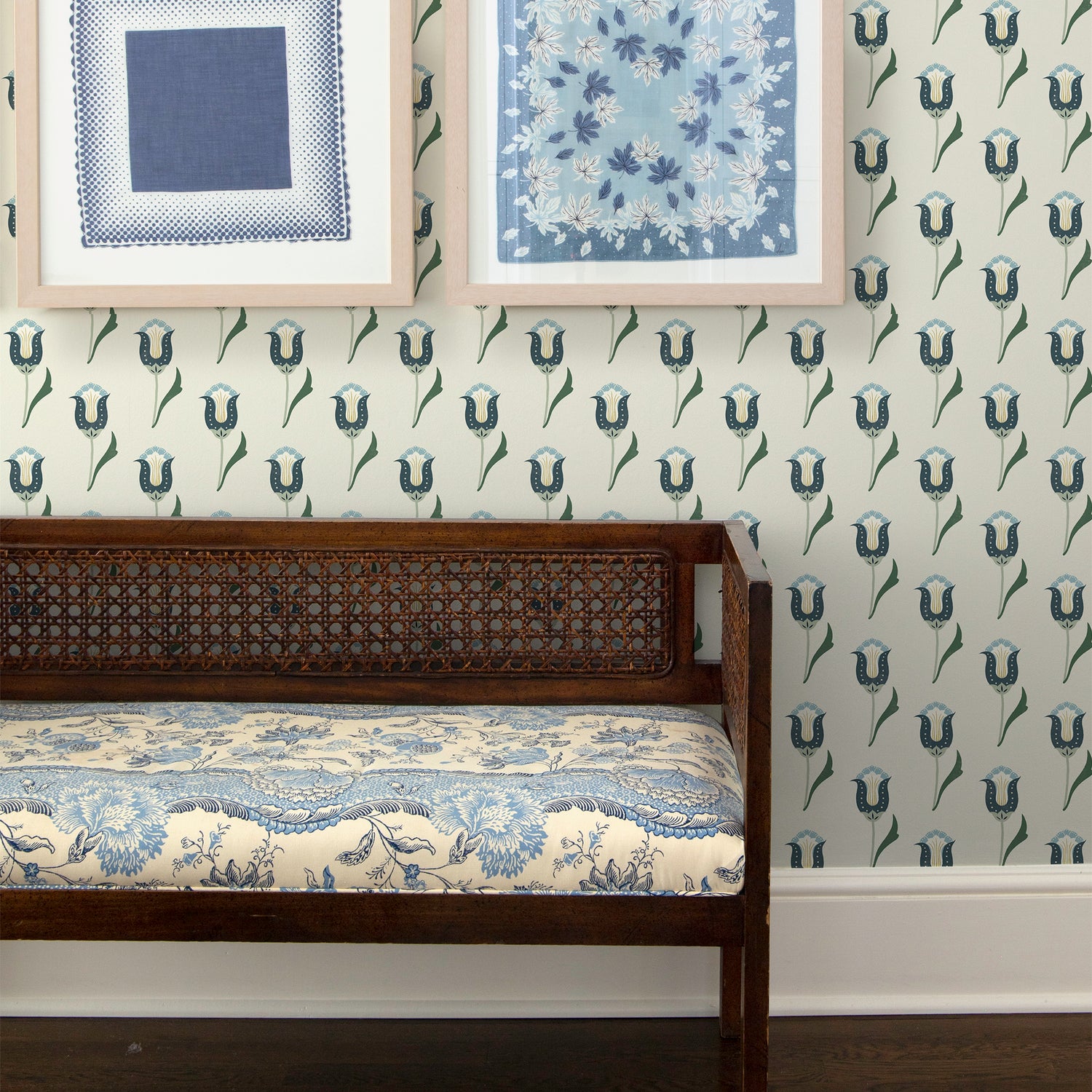 Abstract floral with varying green tones wallpaper on a wall with a brown bench in front with a blue cushion on it and blue framed artwork on the wall