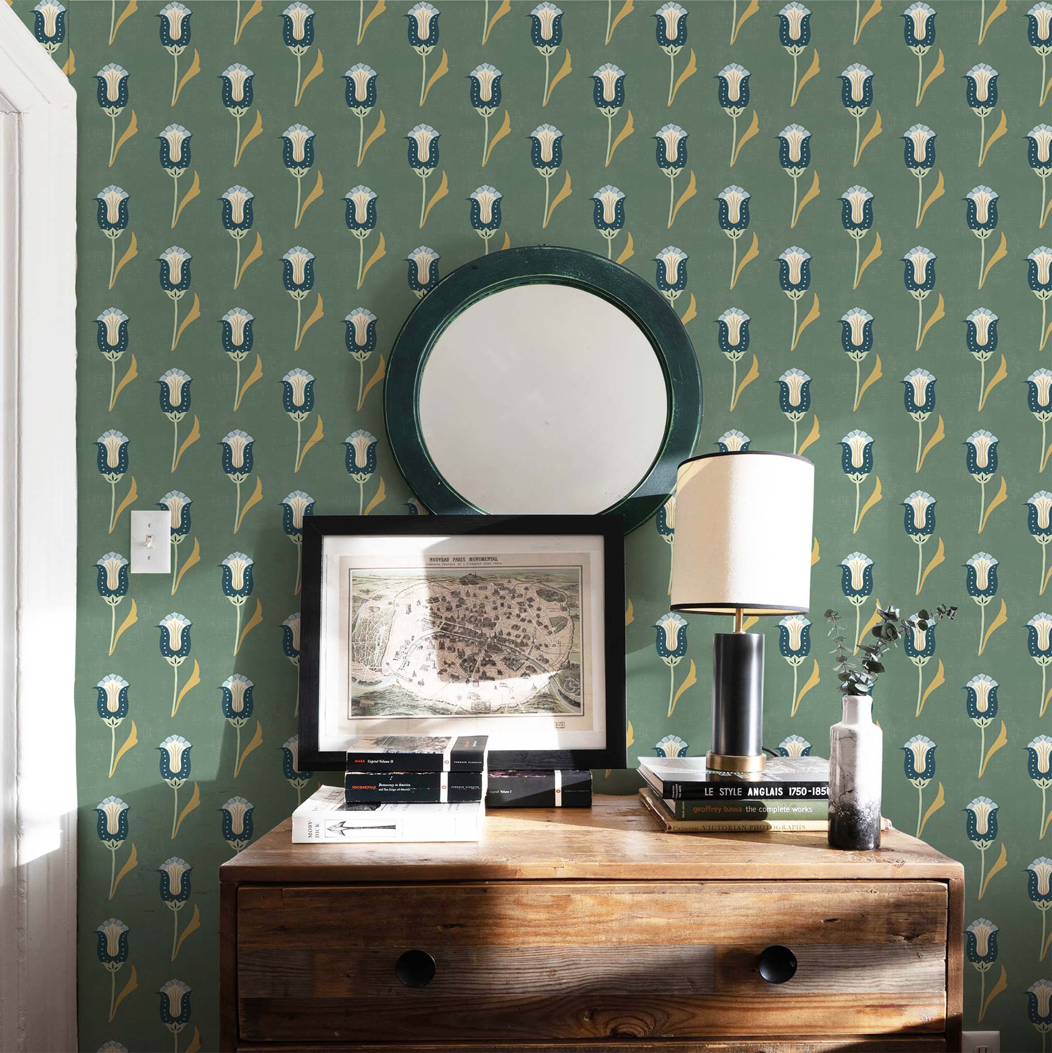 Abstract floral moss green and teal printed wallpaper on a wall with a green mirror hung on the wall and a brown dresser stacked with books