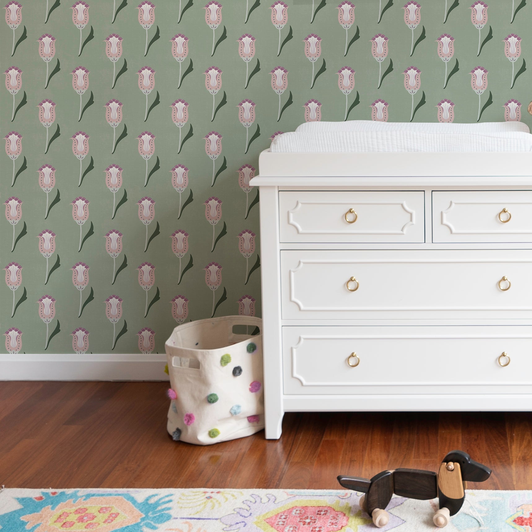 sage green wallpaper with pink and forest green abstract floral pattern on a wall with a white baby changing table in front with drawers and a colorful rug