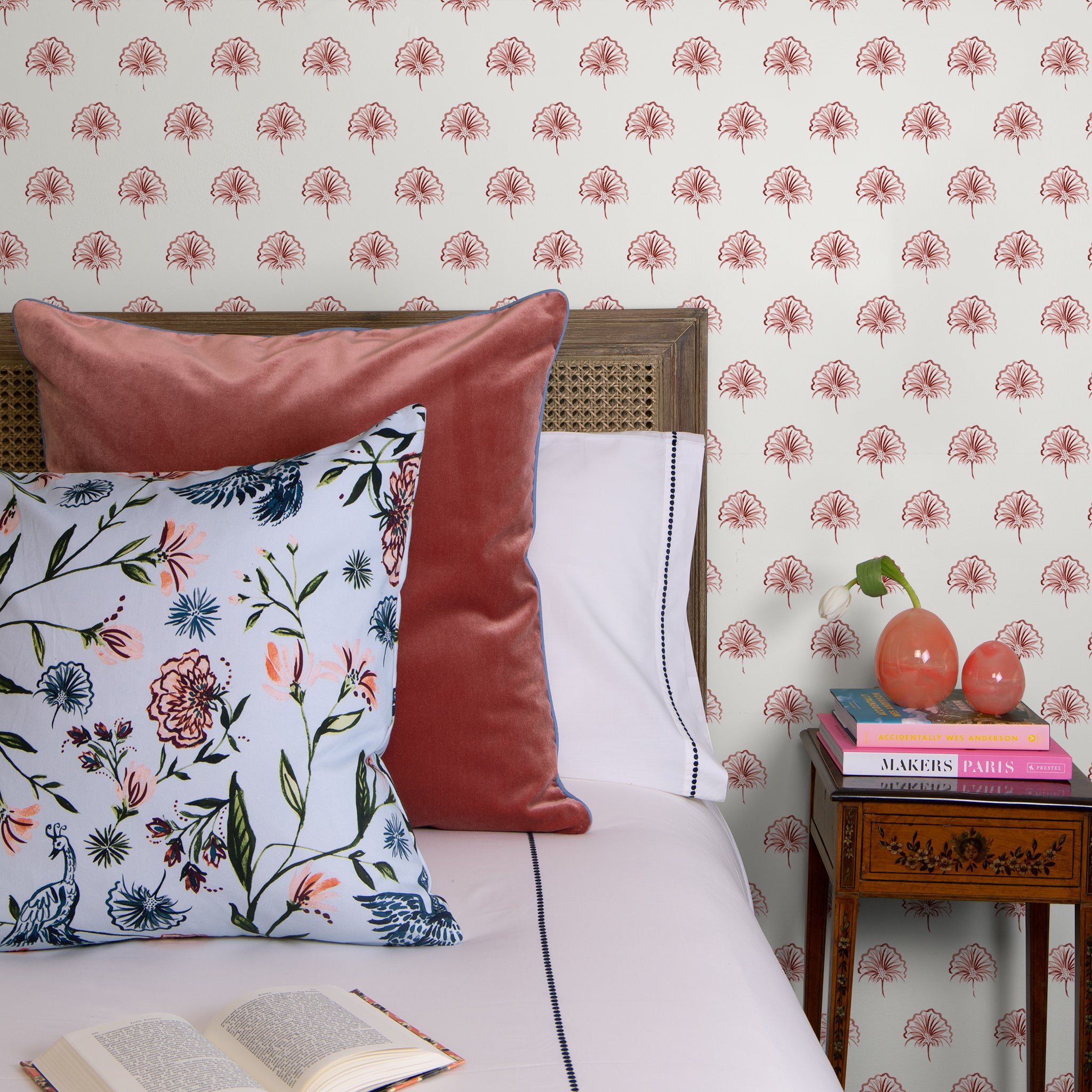 White bed styled with Daphne Powder and Coral Velvet Pillow next to small wooden night stand with books stacked and two crystal decorations on top in front of Rose Floral Printed Wallpaper