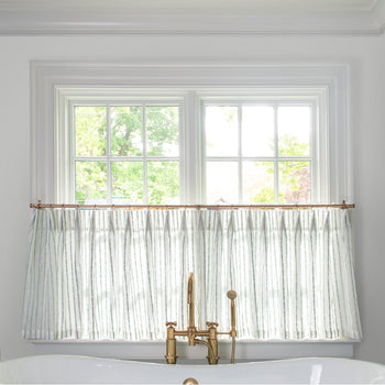 sheer white with mint green embroidered stripe cafe curtain hung in front of an illuminated window behind a white bathtub with a gold faucet 