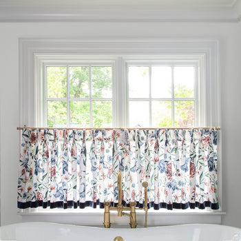 cream chinoiserie cafe curtain hung in front of an illuminated window behind a white bathtub with a gold faucet 