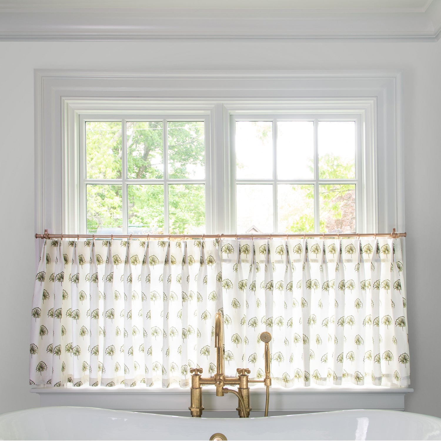 green floral cafe curtain hung in front of an illuminated window behind a white bathtub with a gold faucet 