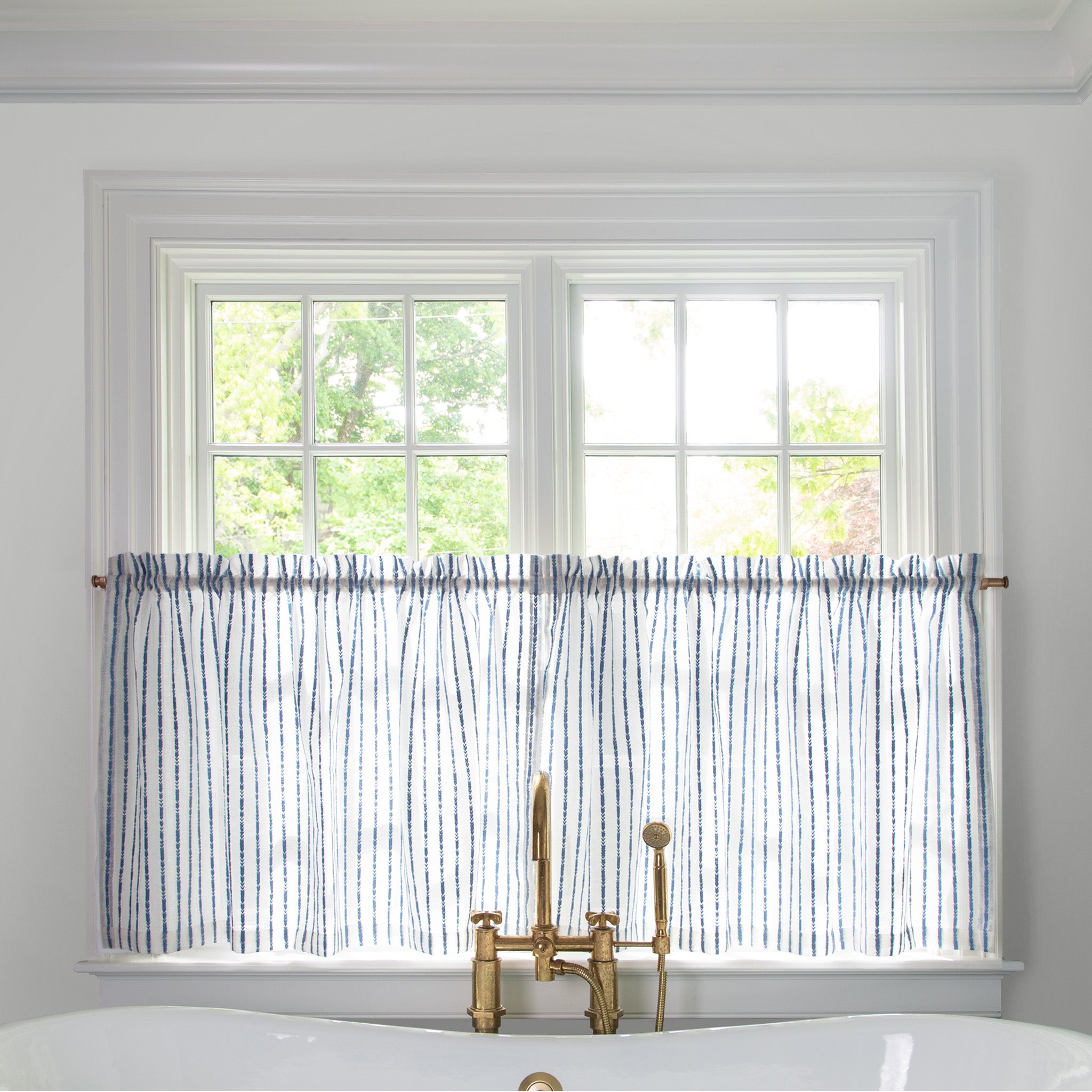 sheer white with embroidered cobalt blue stripe cafe curtain hung in front of an illuminated window behind a white bathtub with a gold faucet 