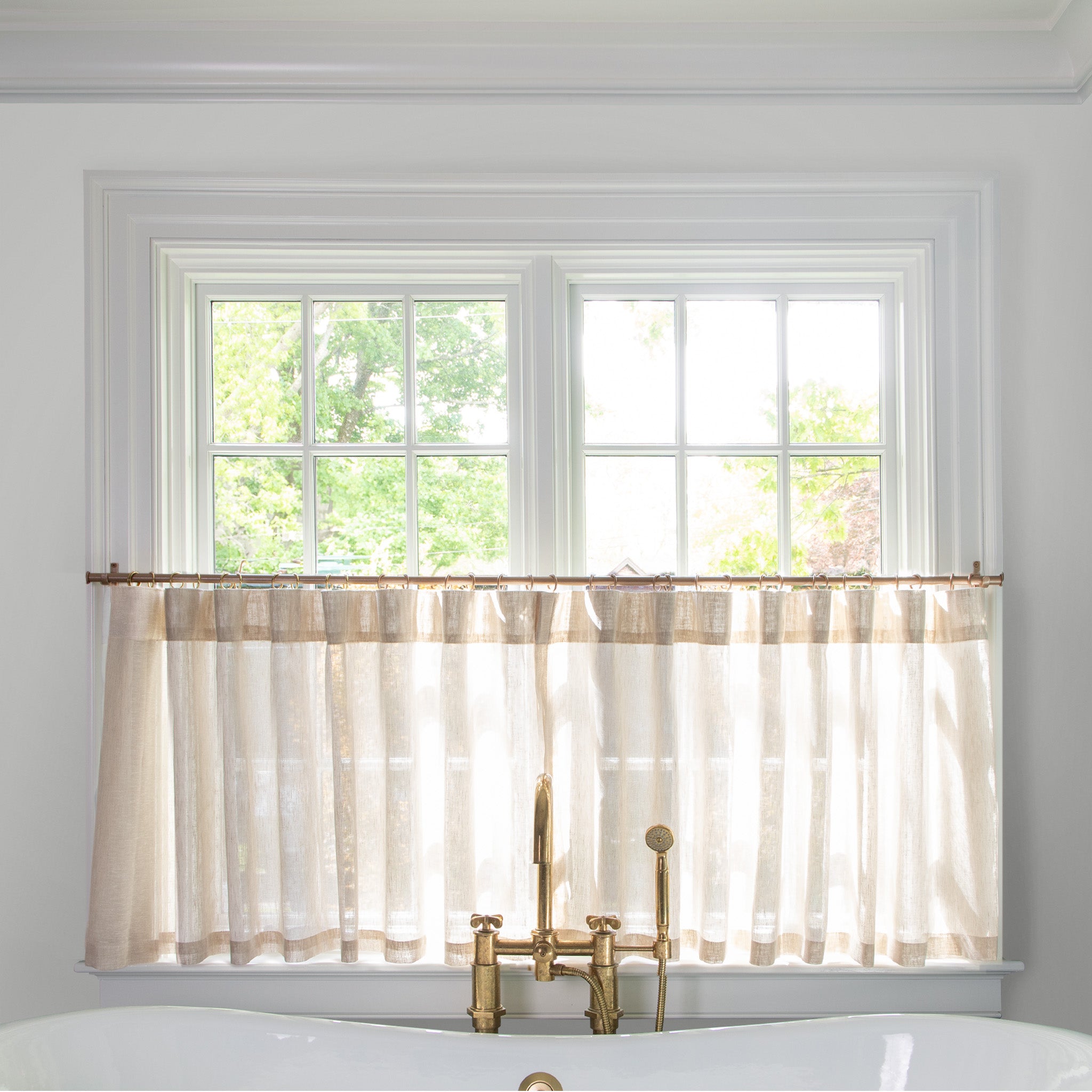 sheer natural brown color cafe curtain hung in front of an illuminated window behind a white bathtub with a gold faucet 