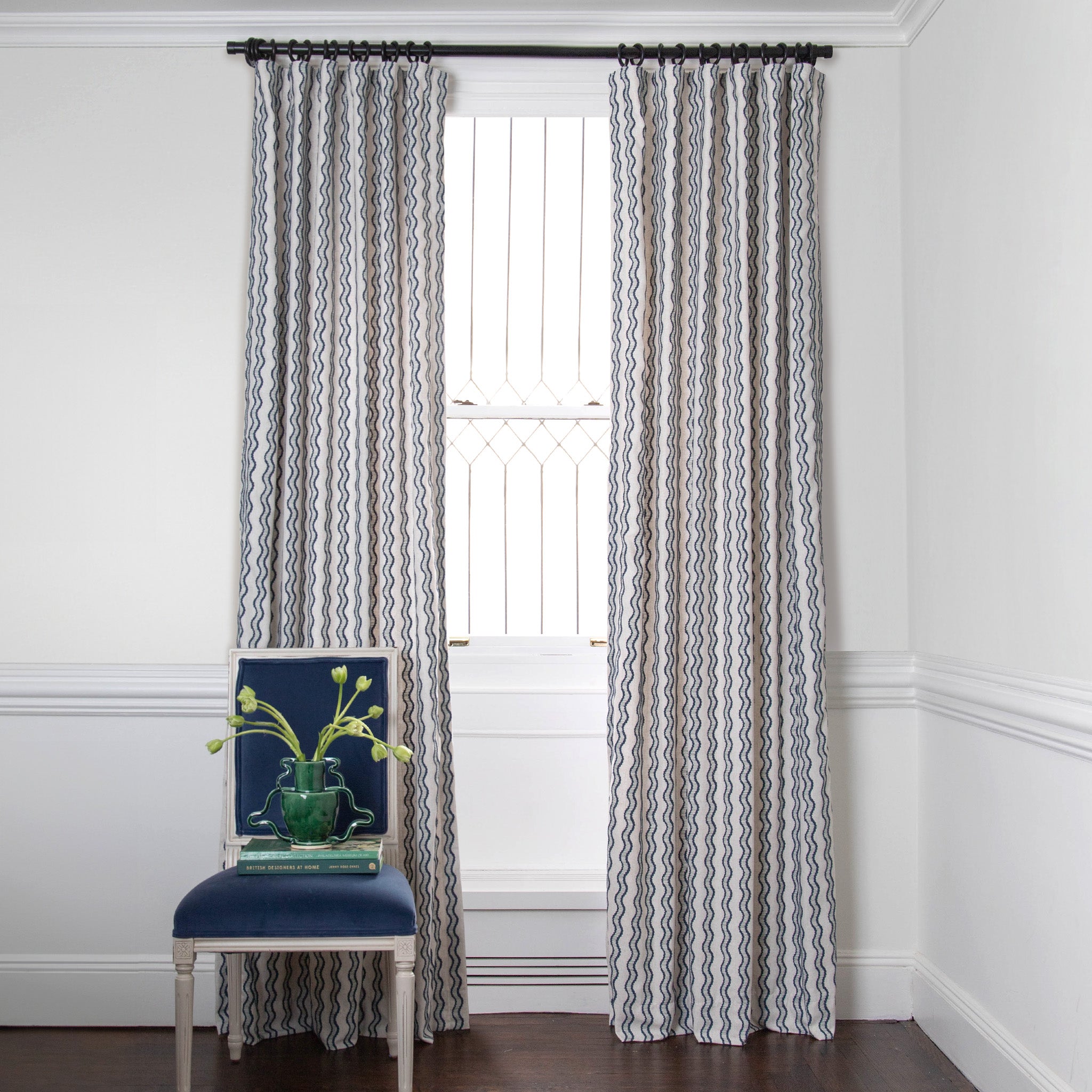 embroidered cream curtains with navy blue wavy lines on a metal rod in front of an illuminated window with a blue chair stacked with books 