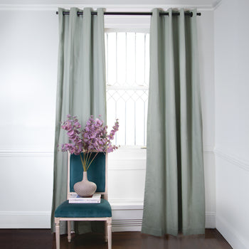 Sage Green Curtains on metal rod in front of an illuminated window with Fern Green Velvet chair with purple flowers in vase on top of stacked books