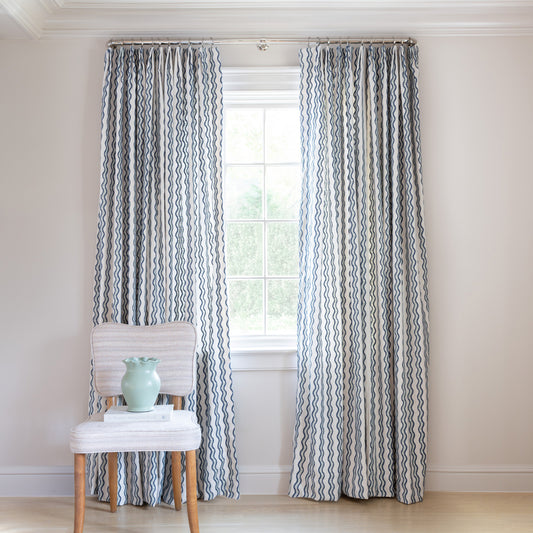 embroidered cream curtains with navy blue wavy lines on a metal rod in front of an illuminated window with a blue chair stacked with books 