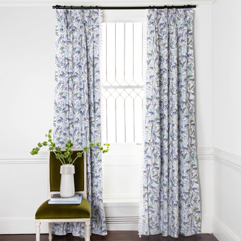 Blue and green floral printed curtains on metal rod in front of an illuminated window with green chair in front stacked with books and a white vase with green flowers