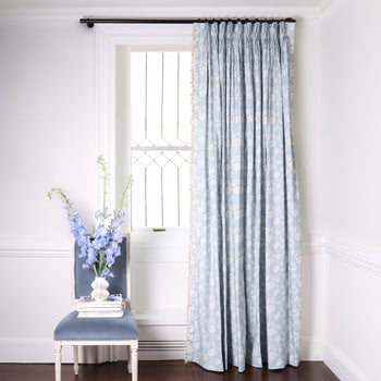 blue floral curtain hung in front of an illuminated window with a blue velvet chair in front of the window with books and flowers on the chair