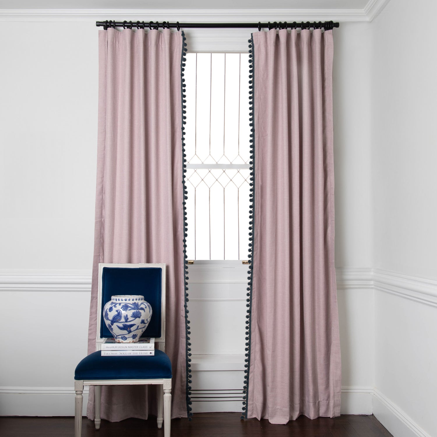 pink curtains with navy blue pom pom trim hung on a black metal rod in front of an illuminated window with a navy blue velvet chair in front stacked with books and a vase 