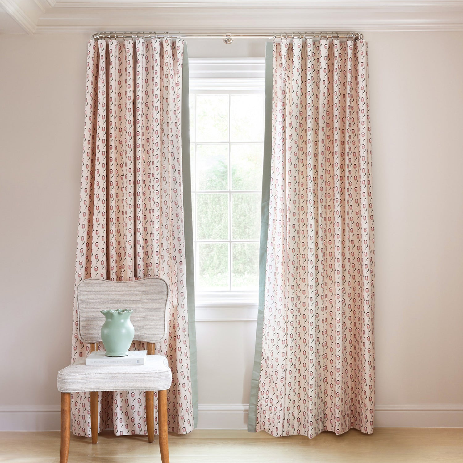 abstract floral pink and green patterned curtains on metal rod in front of an illuminated window with a green chair in front with flowers on it