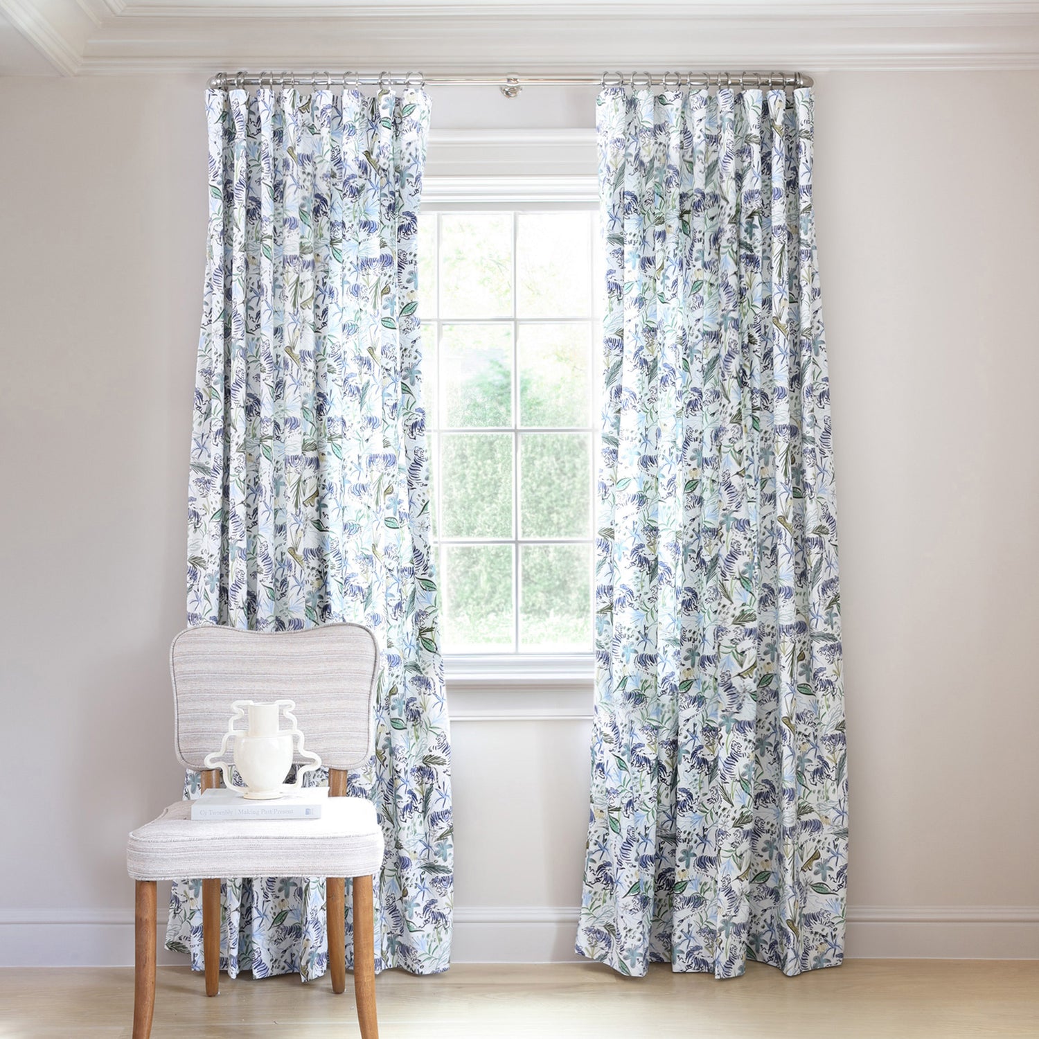 Blue and green floral printed curtains on metal rod in front of an illuminated window with green chair in front stacked with books and a white vase with green flowers