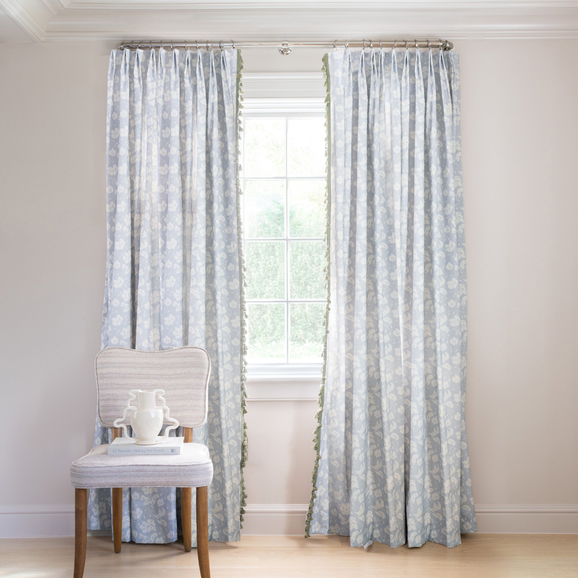 blue floral curtain hung in front of an illuminated window with a blue velvet chair in front of the window with books and flowers on the chair