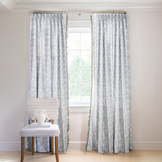 blue floral curtain hung in front of an illuminated window with a blue velvet chair in front of the window with books and flowers on the chair