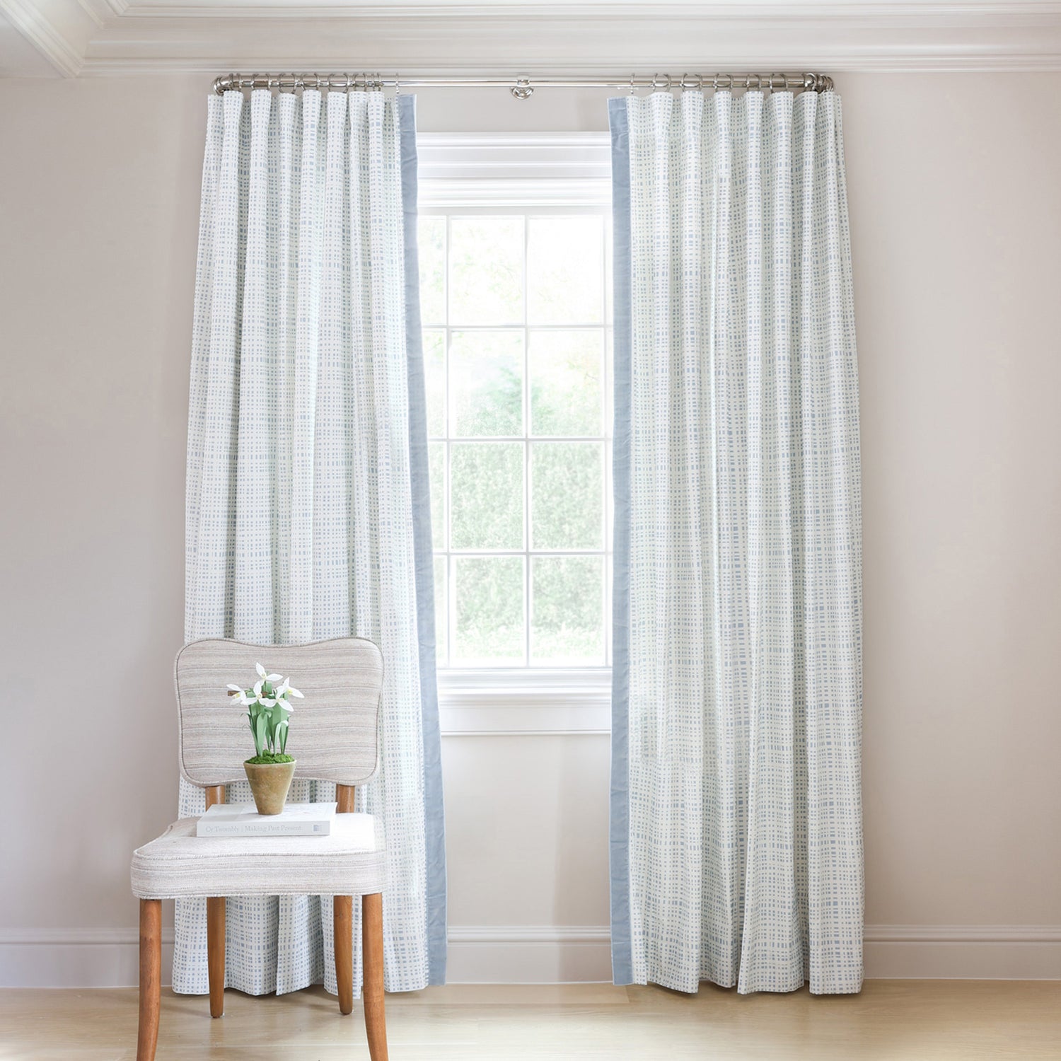 Sky Blue Gingham Printed Curtains on metal rod in front of an illuminated window with Blue Velvet chair with white flowers in blue vase on top of stacked books