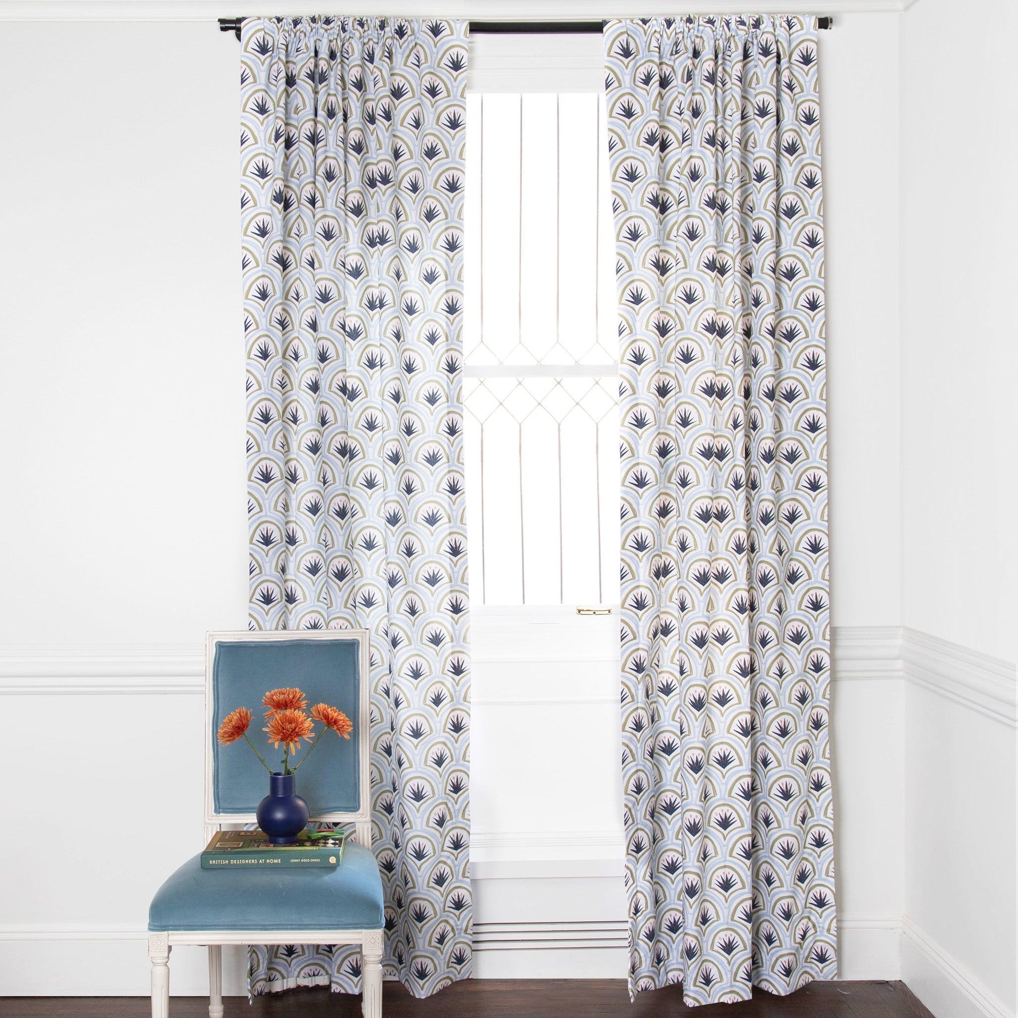 Art Deco Palm Pattern Printed Curtains on white rod in front of an illuminated window with Blue Velvet chair with orange flowers in blue vase on top of green book