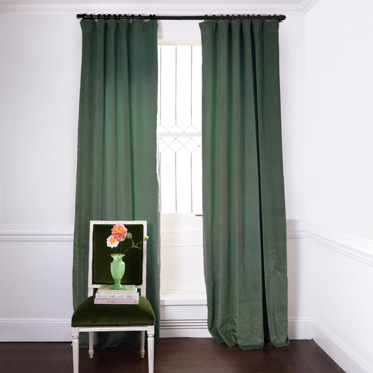 Fern Green Velvet Curtains on black rod in front of an illuminated window with Fern Green Velvet chair with flowers in green vase on top of two stacked books