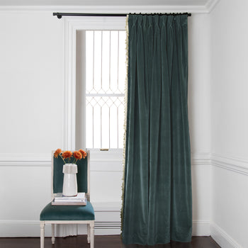 peacock blue velvet curtain on a metal rod in front of an illuminated window with a teal blue chair in front stacked with books