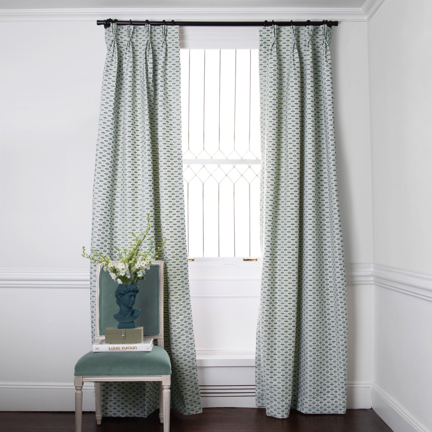 chenille and woven jacquard mint green geometric curtain hung in front of an illuminated window with a teal velvet chair in front of the curtains stacked with books and a bust