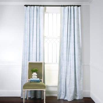 Sky Blue Palm Printed Curtains on metal rod in front of an illuminated window with Moss Green Velvet chair with plant in white vase on top of stacked books