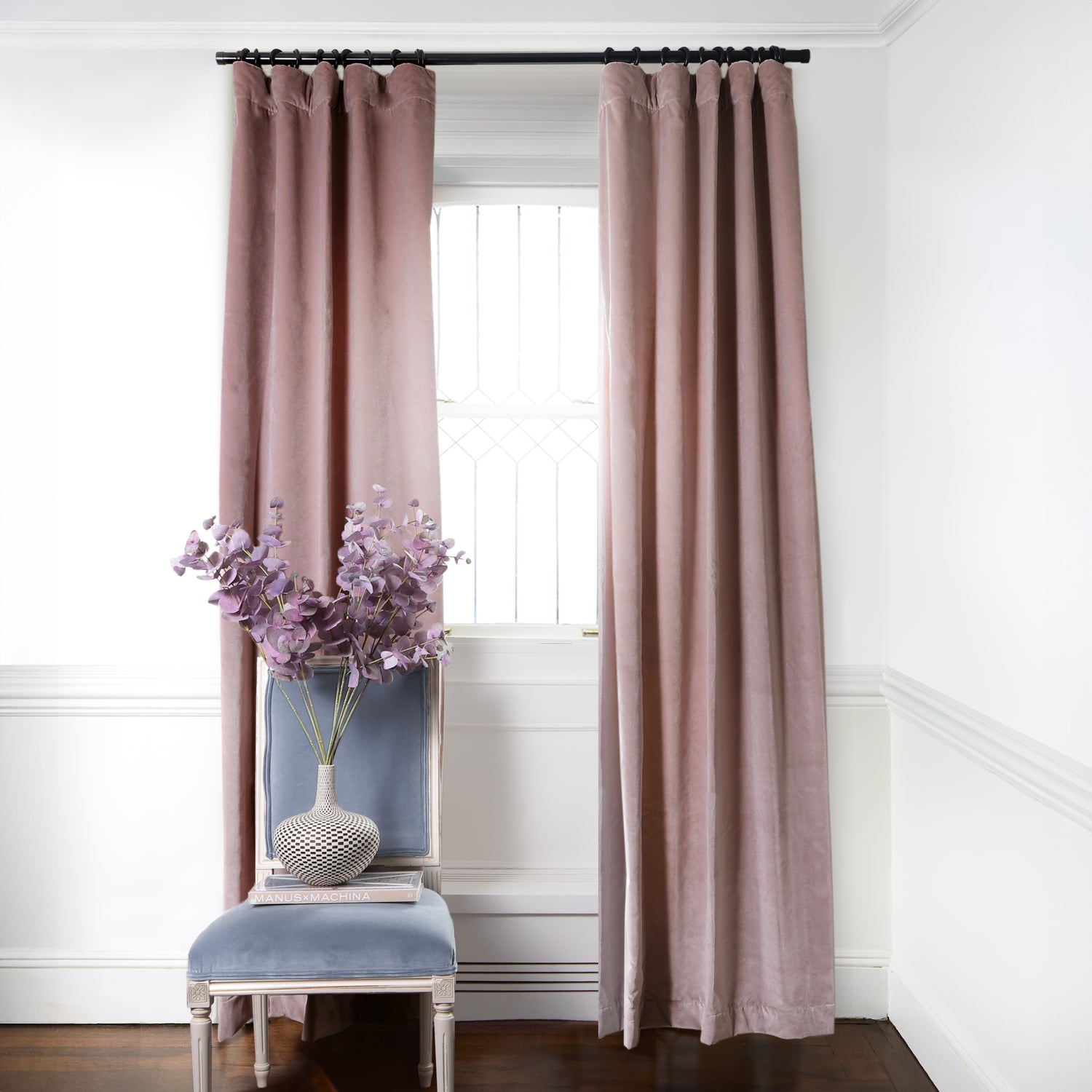 Mauve Velvet Curtains on metal rod in front of an illuminated window with Sky Blue Velvet chair with purple flowers in white and black vase on top book