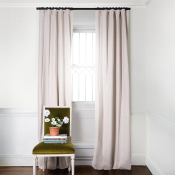 Oat Linen Curtain on metal rod in front of an illuminated window with moss green velvet chair and white flowers in clay pot on stacked books